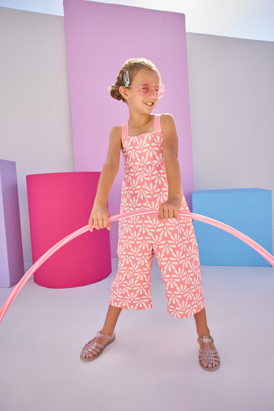 Girls/Tween model can be seen in knit jumpsuit in a salmon/pink and white floral print.This jumpsuit has adjustable woven straps and pockets for easy relaxed fit.