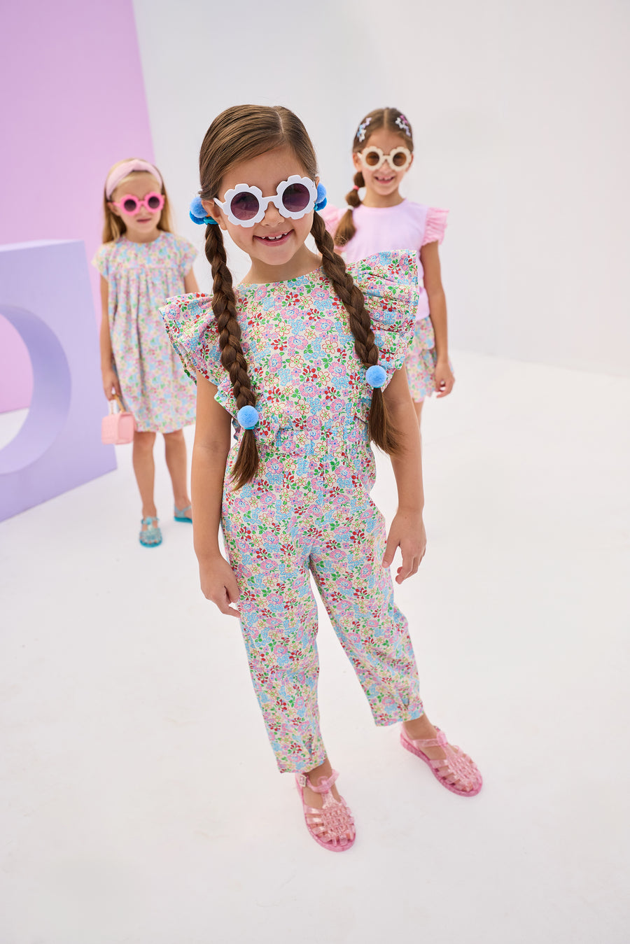 Girls/Tween model in front can be seen wearing a woven jumpsuit that features ruffle detail along the front/sleeves, pockets, and a front tie with an open back. Jumpsuit has a beautiful pale yellow/pink/green/blue floral pattern and is a lightweight woven material for ultimate comfort. 