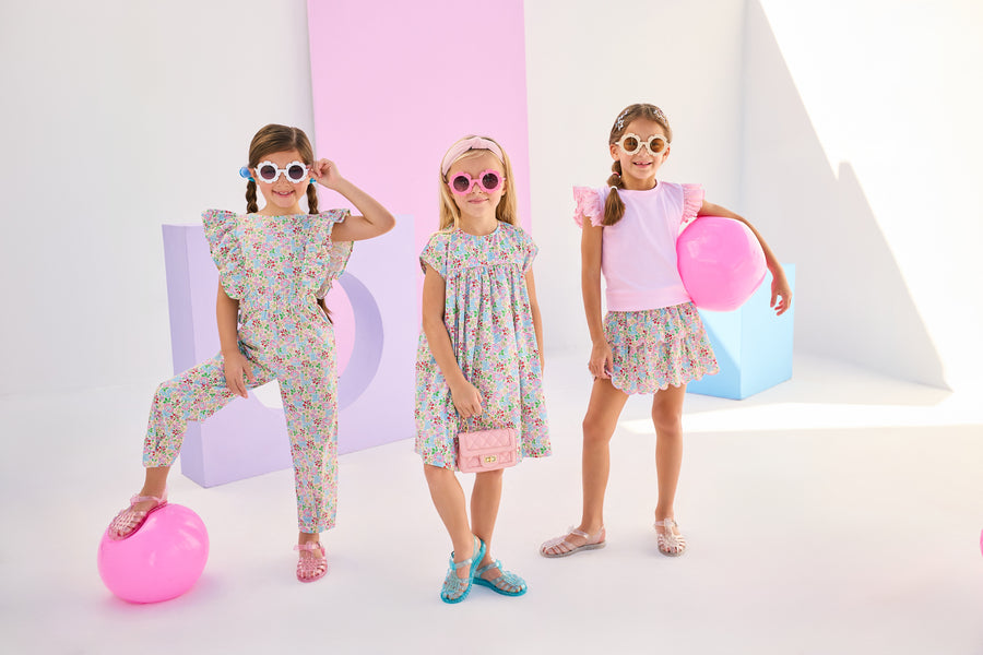 Girls/Tween model on left can be seen wearing a woven jumpsuit that features ruffle detail along the front/sleeves, pockets, and a front tie with an open back. Jumpsuit has a beautiful pale yellow/pink/green/blue floral pattern and is a lightweight woven material for ultimate comfort. Model in the center can be seen in the same floral in a woven dress, and model on right can be seen in the same print with our tiered skort paired back with a light pink eyelet tank. 