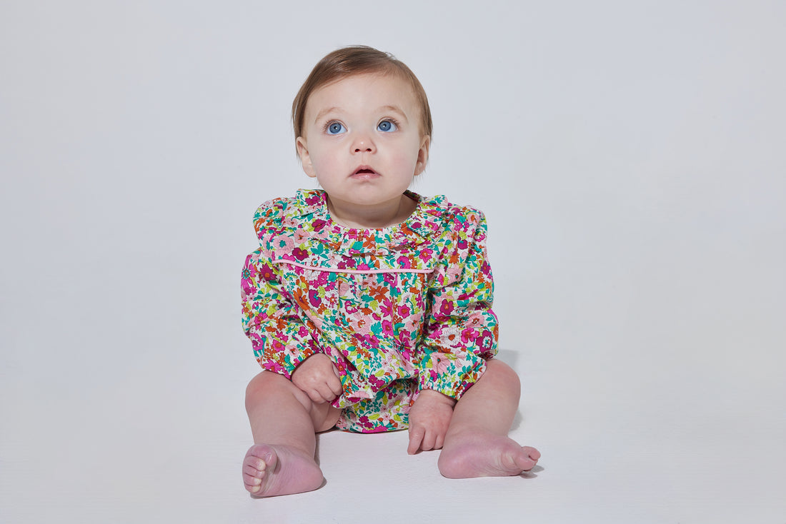 Babygirl long sleeve bubble with elastic wristband featuring a ruffle neckline and button closures along the back and stride in a vibrant magenta/pink/orange/green floral print.