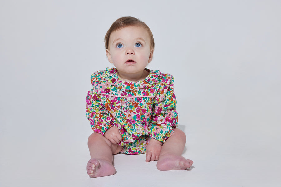 Babygirl long sleeve bubble with elastic wristband featuring a ruffle neckline and button closures along the back and stride in a vibrant magenta/pink/orange/green floral print.