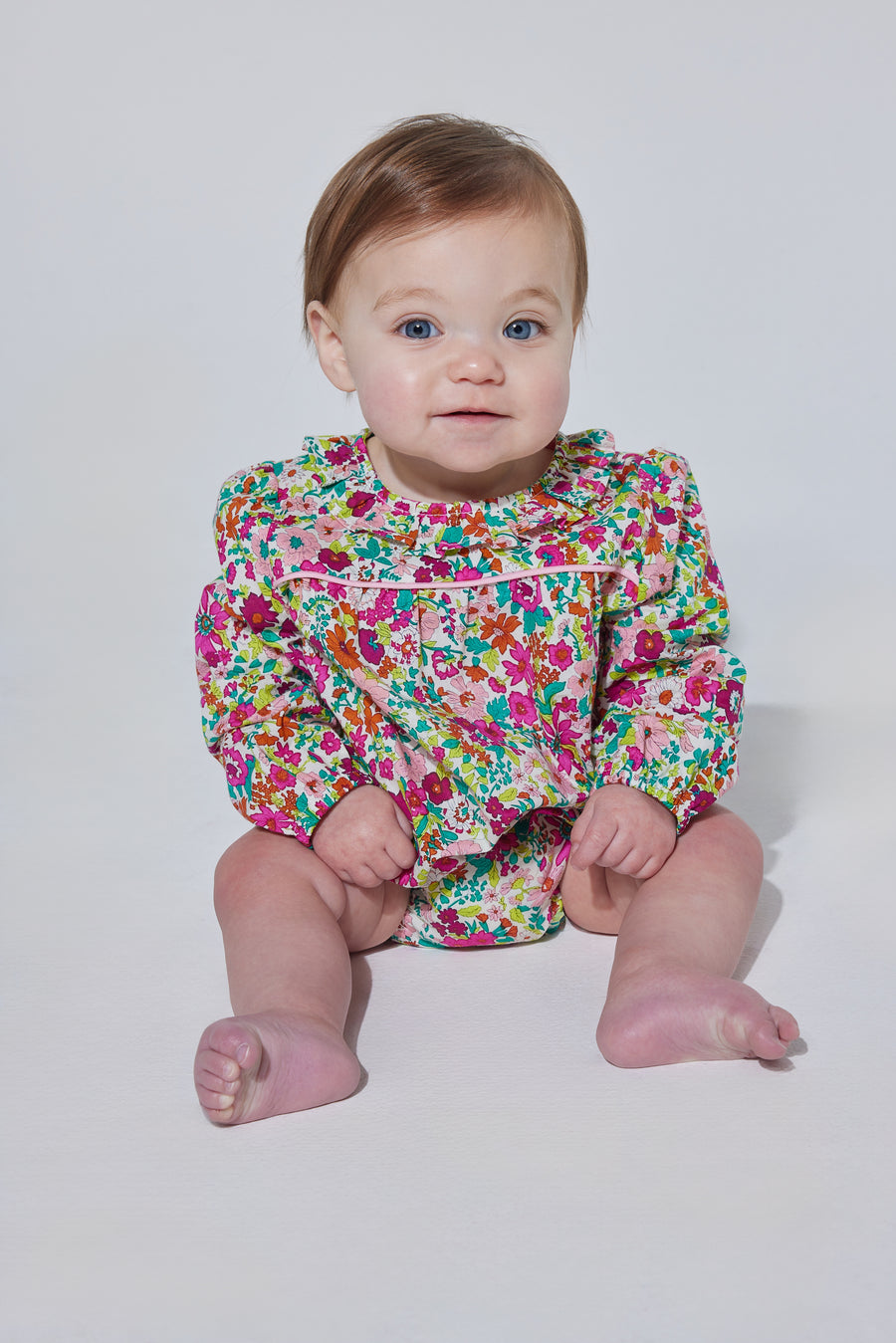 Babygirl long sleeve bubble with elastic wristband featuring a ruffle neckline and button closures along the back and stride in a vibrant magenta/pink/orange/green floral print.