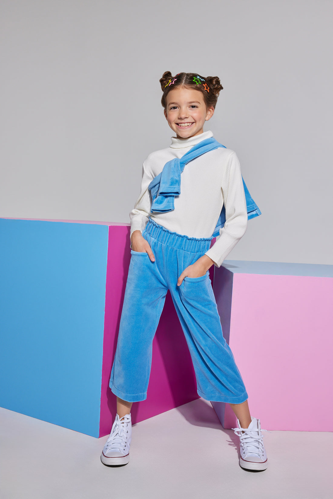 Tween cropped French blue velour pants that have pockets on front and elastic high waisted waistband which can be seen paired with an ivory long sleeve turtleneck and polo pullover thrown over the shoulder.