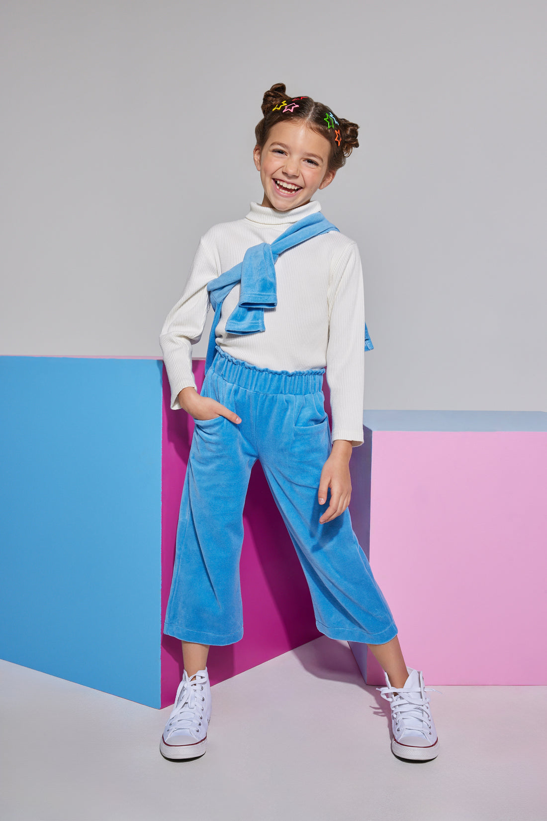 Tween cropped French blue velour pants that have pockets on front and elastic high waisted waistband which can be seen paired with an ivory long sleeve turtleneck and polo pullover thrown over the shoulder.