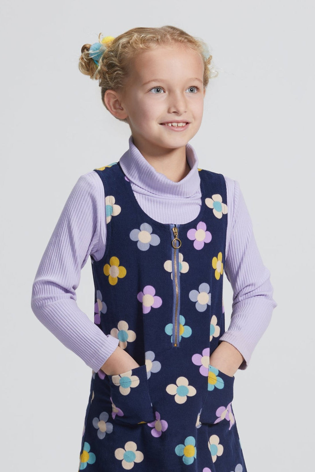 Girls/Tween jumper in a lightweight corduroy material. Jumper has a zipper down the front, pockets, and can be seen in a purple/blue/yellow floral arrangement on a navy background. Perfect as a layering piece, as seen model is wearing a ribbed turtleneck in lilac which pulls back in nicely with the jumper.